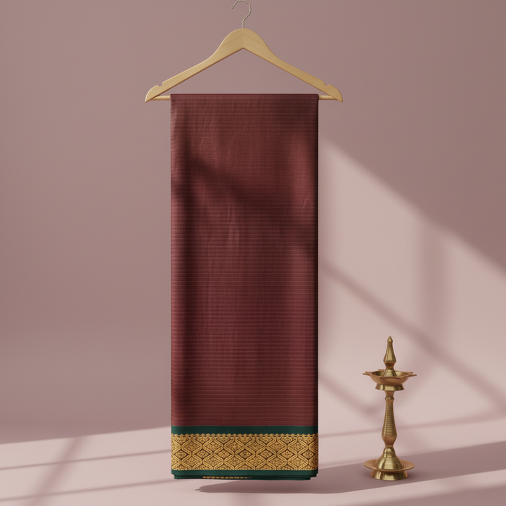 Burgundy striped cotton saree with a forest-green antique-gold border hanging on a wooden hanger against a pastel background with a brass diya