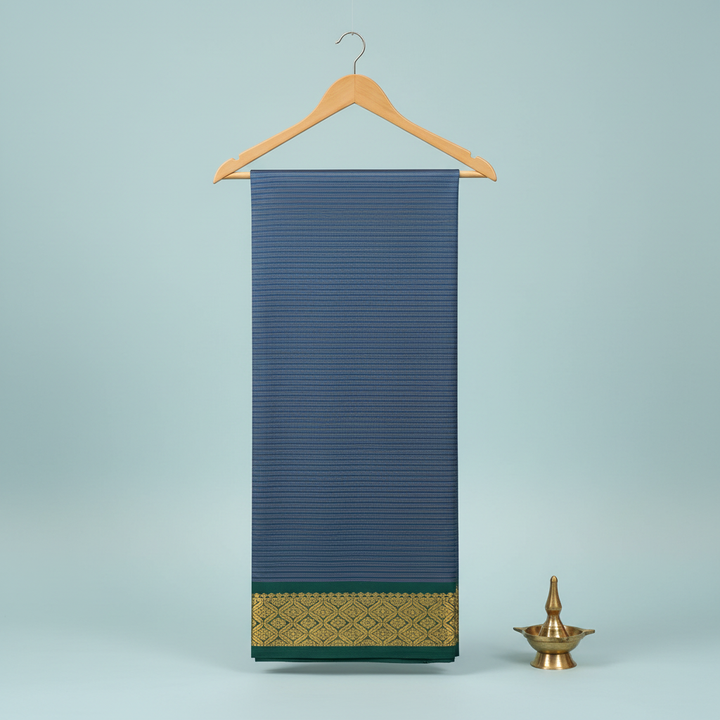 Steel-blue striped cotton saree with a forest-green antique-gold border hanging on a wooden hanger against a pastel background with a brass diya