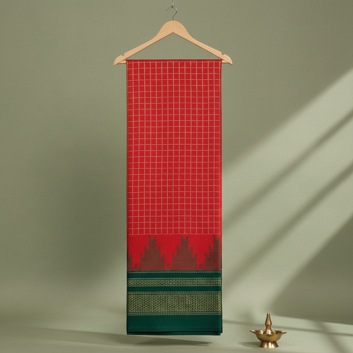 Red and scarlet checked cotton saree with a maroon jagged motif band and dark green patterned border hanging on a wooden hanger against a pastel background with a brass diya