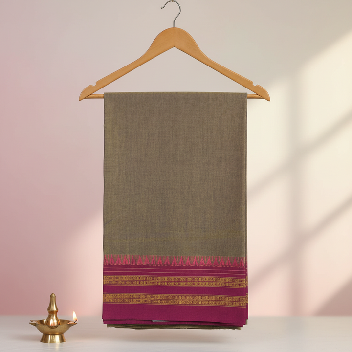 Grey cotton saree with magenta and gold zari border hung on a wooden hanger against a pastel background with a brass lamp