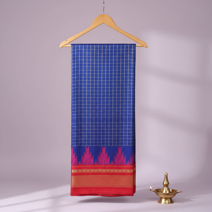 Royal-blue checked cotton saree with a magenta motif band and vivid red-gold border hanging on a wooden hanger against a pastel background with a brass diya