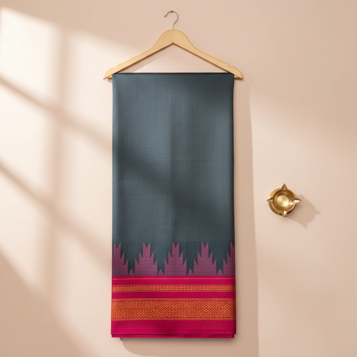 Charcoal-grey cotton saree with a magenta motif band and crimson-gold border hanging on a wooden hanger against a pastel background with a brass diya