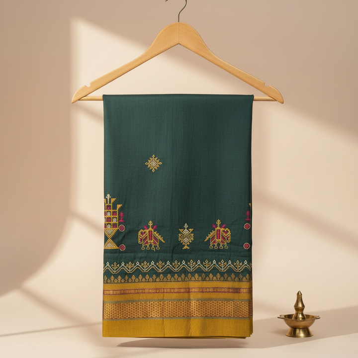Dark green cotton Kasuti saree with chariot and peacock embroidery and a mustard-gold zari border, hung on a wooden hanger against a pastel background with a brass lamp