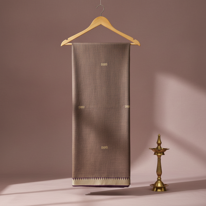 Pale brown cotton saree with small antique-gold motifs and a contrasting ivory-gold geometric border hanging on a wooden hanger against a pastel background with a brass diya