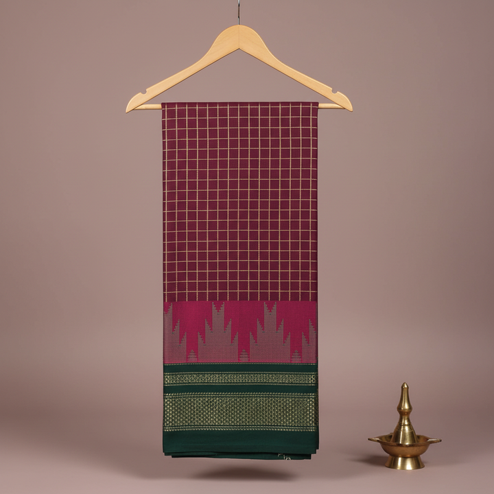 Burgundy and plum checked cotton saree with a magenta motif band and dark green patterned border hanging on a wooden hanger against a pastel background with a brass diya